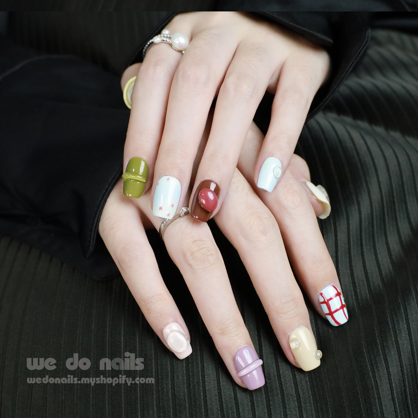 Close-up of colorful hands against a dark background, featuring We Do Nails Cute Abstract Art Press-On Nails with vibrant patterns like stripes and polka dots. Some nails have 3D finishes and embellishments, complemented by several rings on the persons fingers.