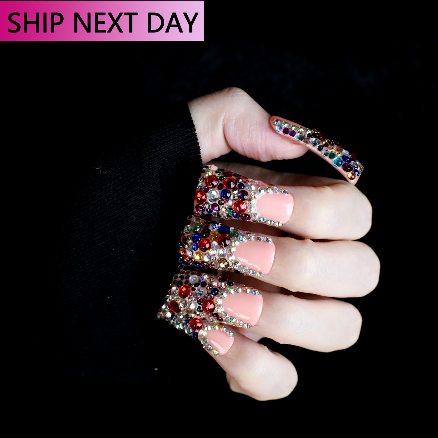 A hand flaunts Dazzling Long Duck Press-On Nails from We Do Nails, featuring a nude pink base with multicolored rhinestones against a black background. The SHIP NEXT DAY label is clearly visible in the top left corner.