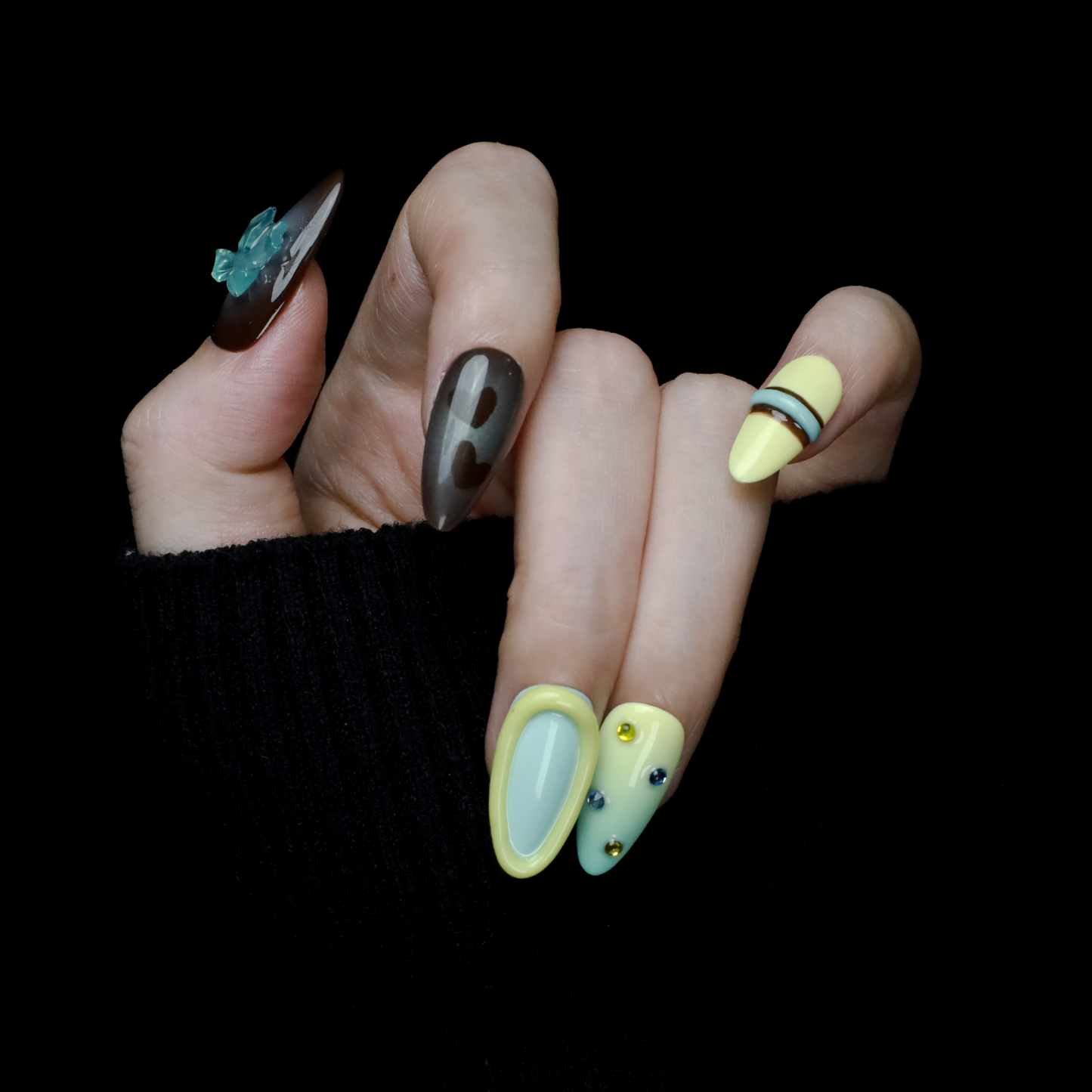 A hand showcases Elegant Edge Press-On Nails by We Do Nails from the Chic Charm Collection against a black background, with designs in brown and blue gem, camo, light yellow with stripes, pale blue with rhinestones, and yellow bordered in lime green; a sleek black sleeve adorns the wrist.