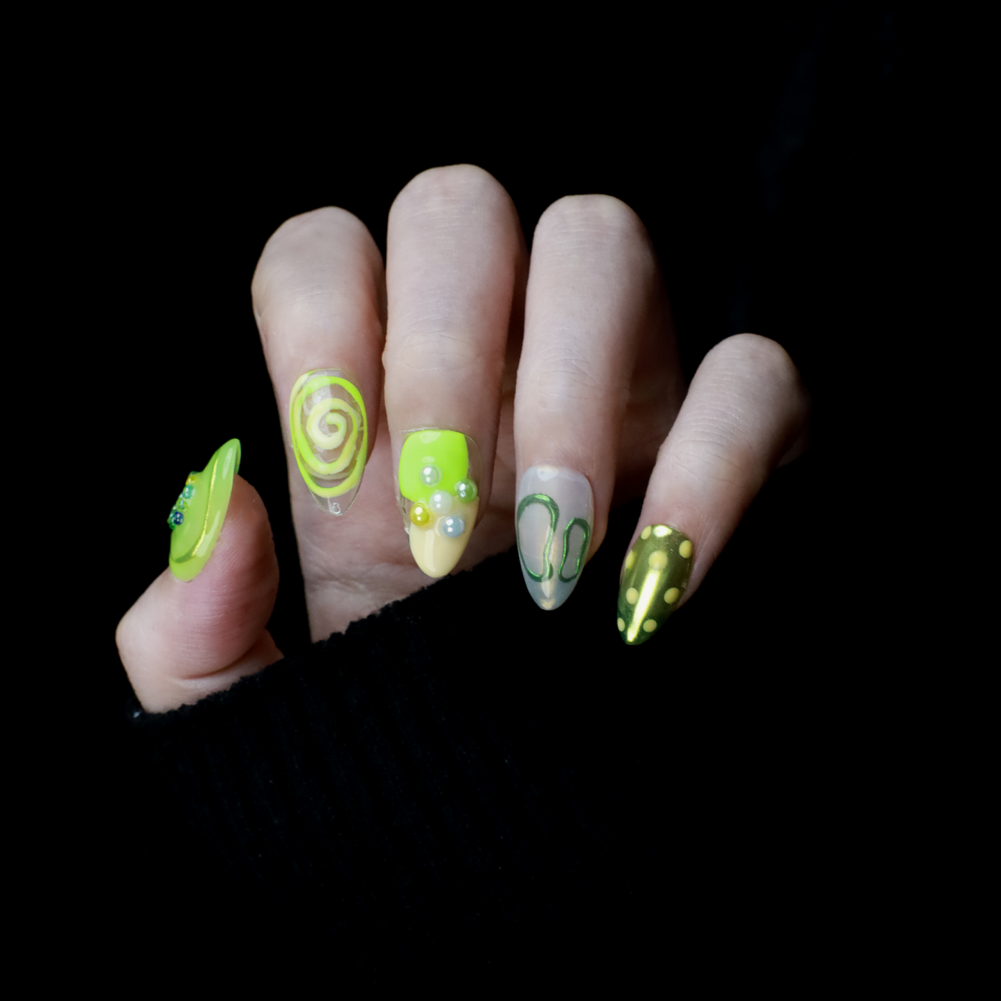 A hand holding a black mug flaunts vibrant We Do Nails Neon Slime Fantasy Press-On Nails, featuring abstract lime green, yellow, and black patterns. Spirals, dots with bead accents, and 3D smiley faces create playful art against the dark backdrop.