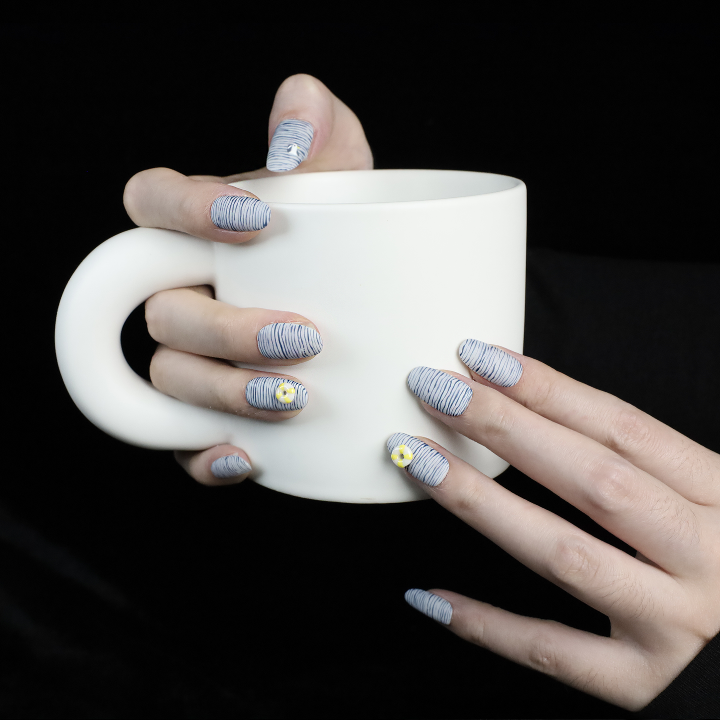 A hand with long nails showcases We Do Nails Nautical Navy Blue Stripes press-on nails, featuring a nautical blue and black wavy design. One nail is adorned with a 3D yellow flower, another with a small gem. A black sleeve adds elegance to the hand-painted look.