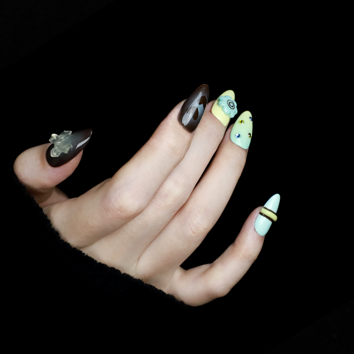 A hand showcases Elegant Edge Press-On Nails by We Do Nails from the Chic Charm Collection against a black background, with designs in brown and blue gem, camo, light yellow with stripes, pale blue with rhinestones, and yellow bordered in lime green; a sleek black sleeve adorns the wrist.