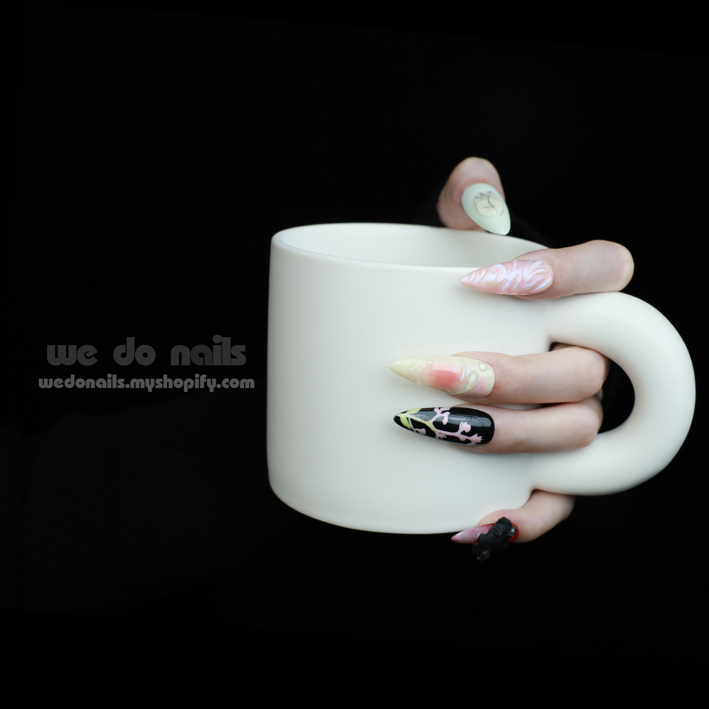 A hand holds a white mug, displaying We Do Nails Abstract Floral Design Press-On Nails, featuring intricate floral patterns in vibrant colors against a dark backdrop. Text: We Do Nails wedonails.myshopify.com.