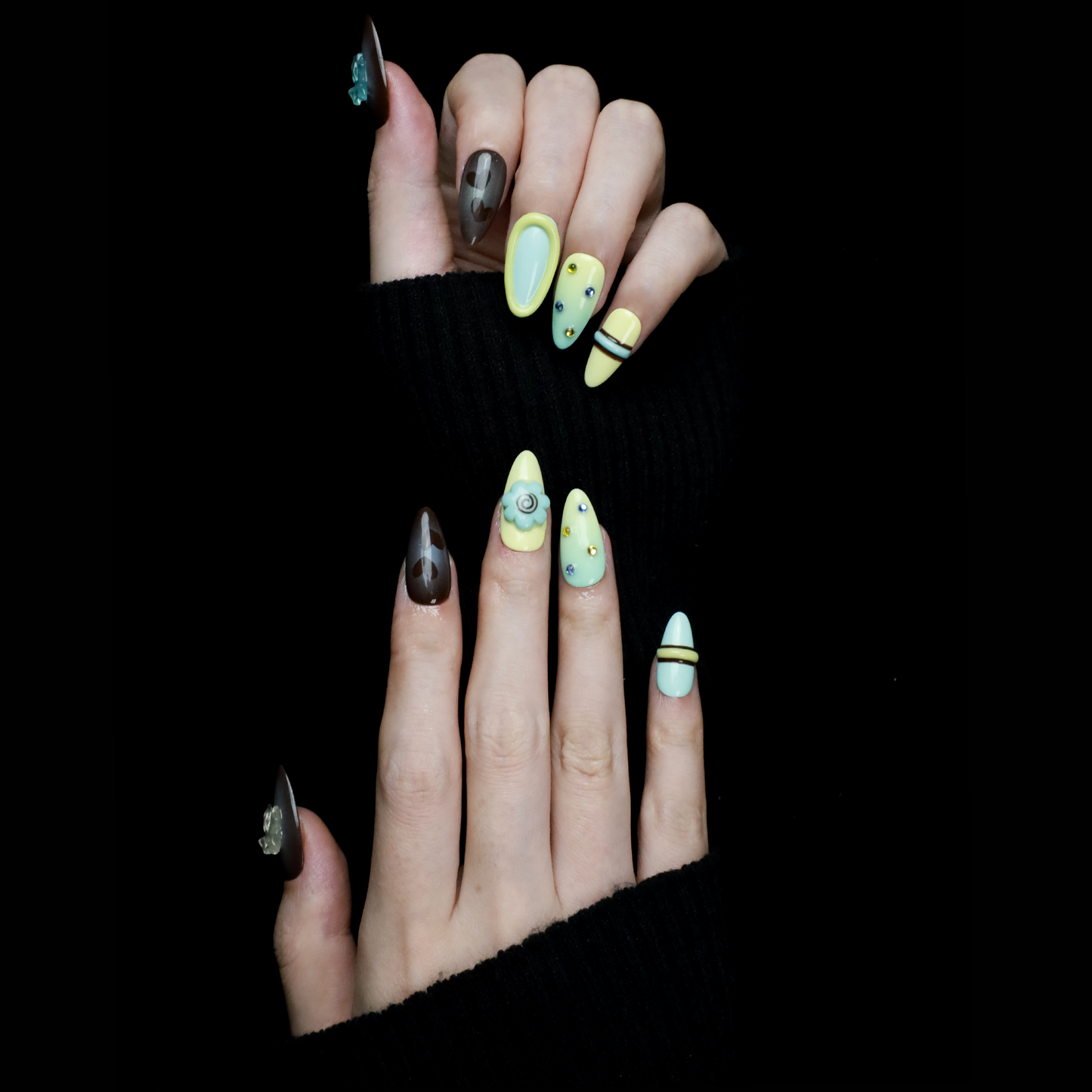 A hand showcases Elegant Edge Press-On Nails by We Do Nails from the Chic Charm Collection against a black background, with designs in brown and blue gem, camo, light yellow with stripes, pale blue with rhinestones, and yellow bordered in lime green; a sleek black sleeve adorns the wrist.