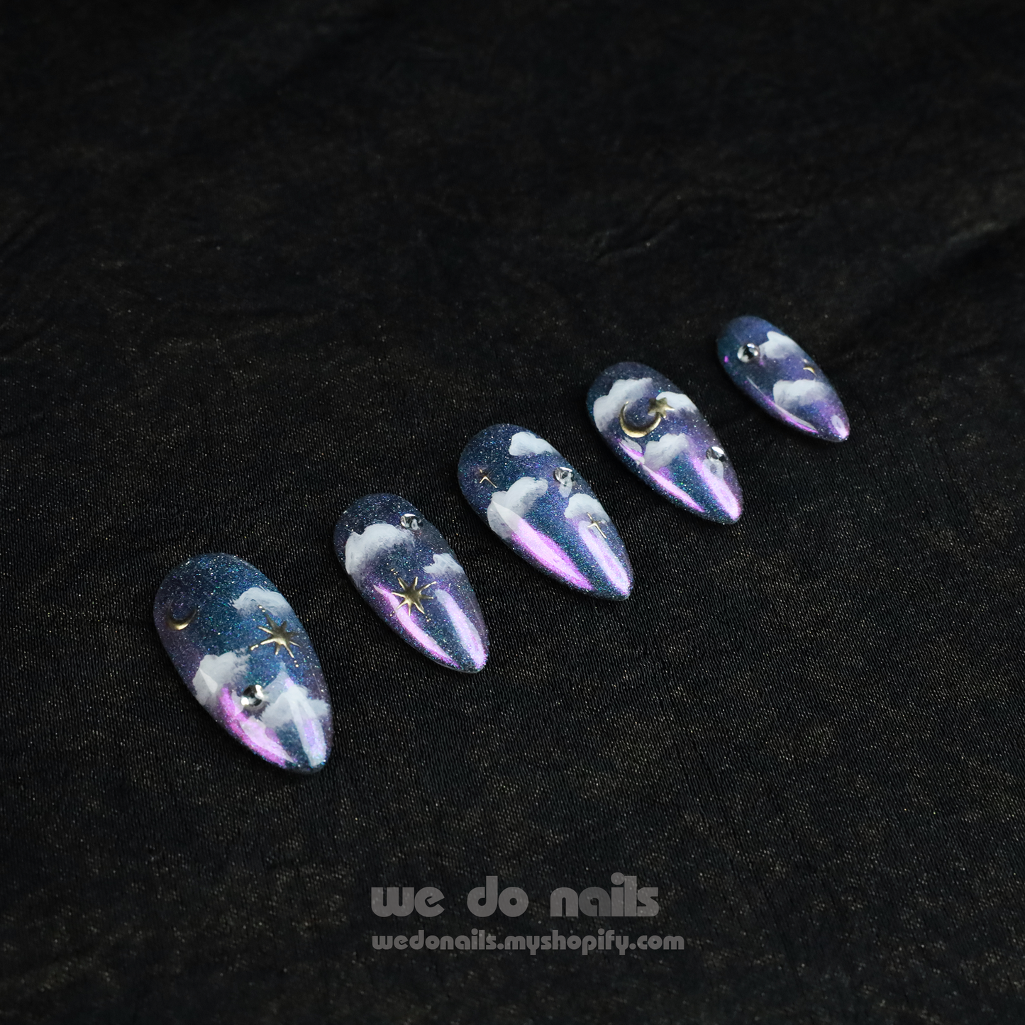 A hand showcases Starry Night Press-On Nails with long, glittery tips featuring a cloud and starry night design adorned with small gems. The person wears a black sleeve. Text below reads: We Do Nails with a website link.