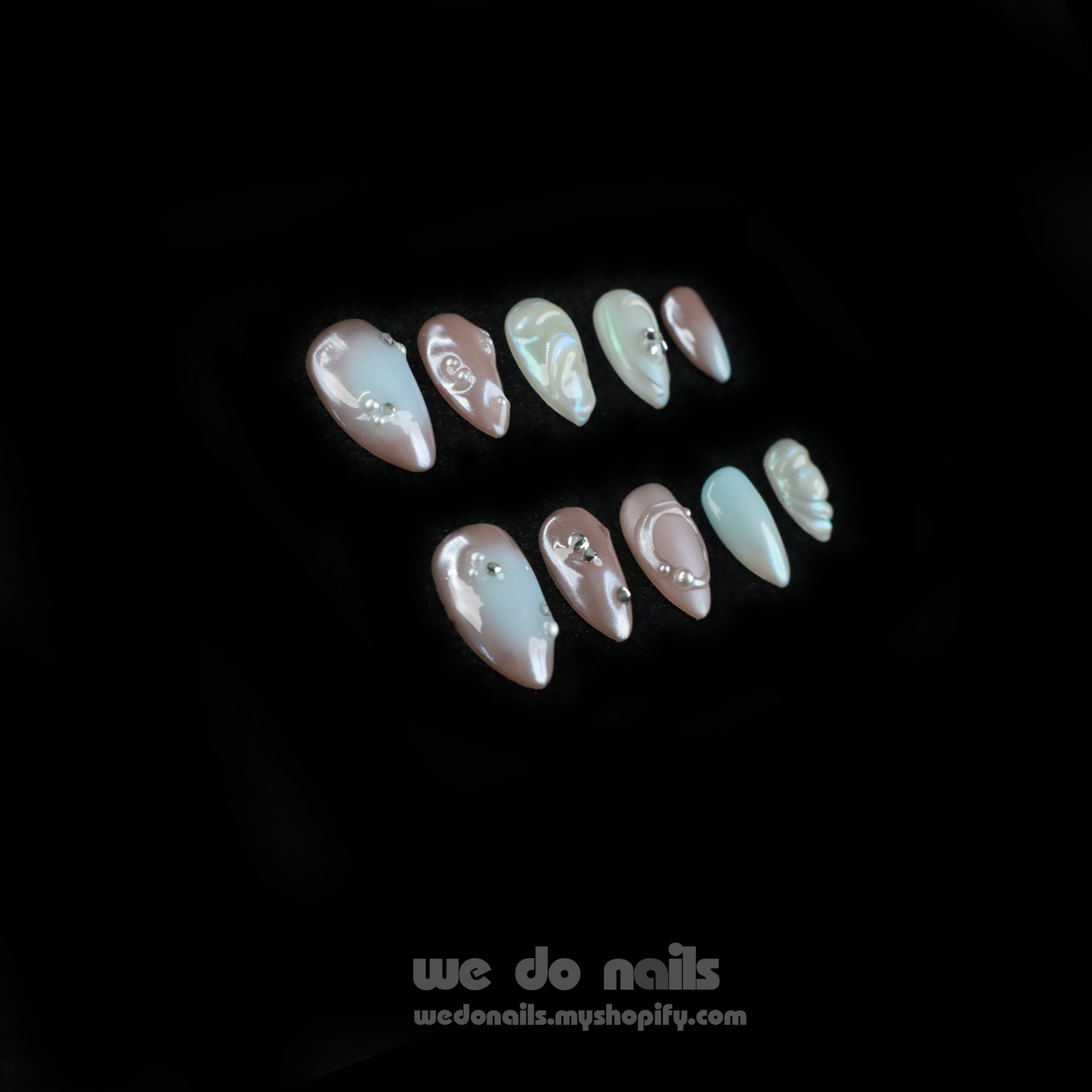Close-up of hands with Pearl Dreams Press-On Nails by We Do Nails, showcasing manicured nails in small patterns and neutral colors. Subtle pearlescent hues add elegance. Fingers are gently intertwined, wearing a black long-sleeve sweater. We Do Nails text visible on the left.