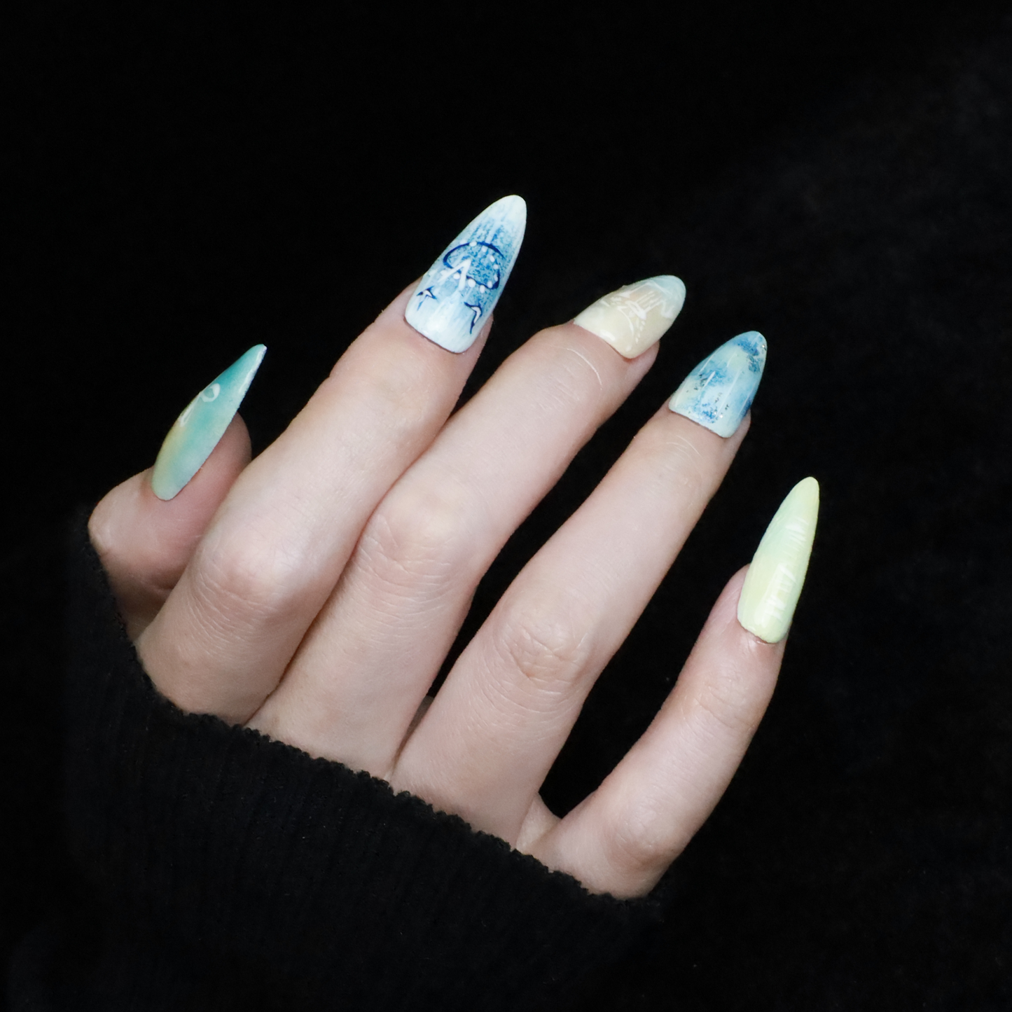 A hand flaunts the elegant Ocean-Inspired Press-On Nails by We Do Nails, featuring long, pointed shapes with ocean-themed designs. The set showcases ocean breeze colors, seashell art, and wave patterns against a dark background to highlight its intricate beauty.