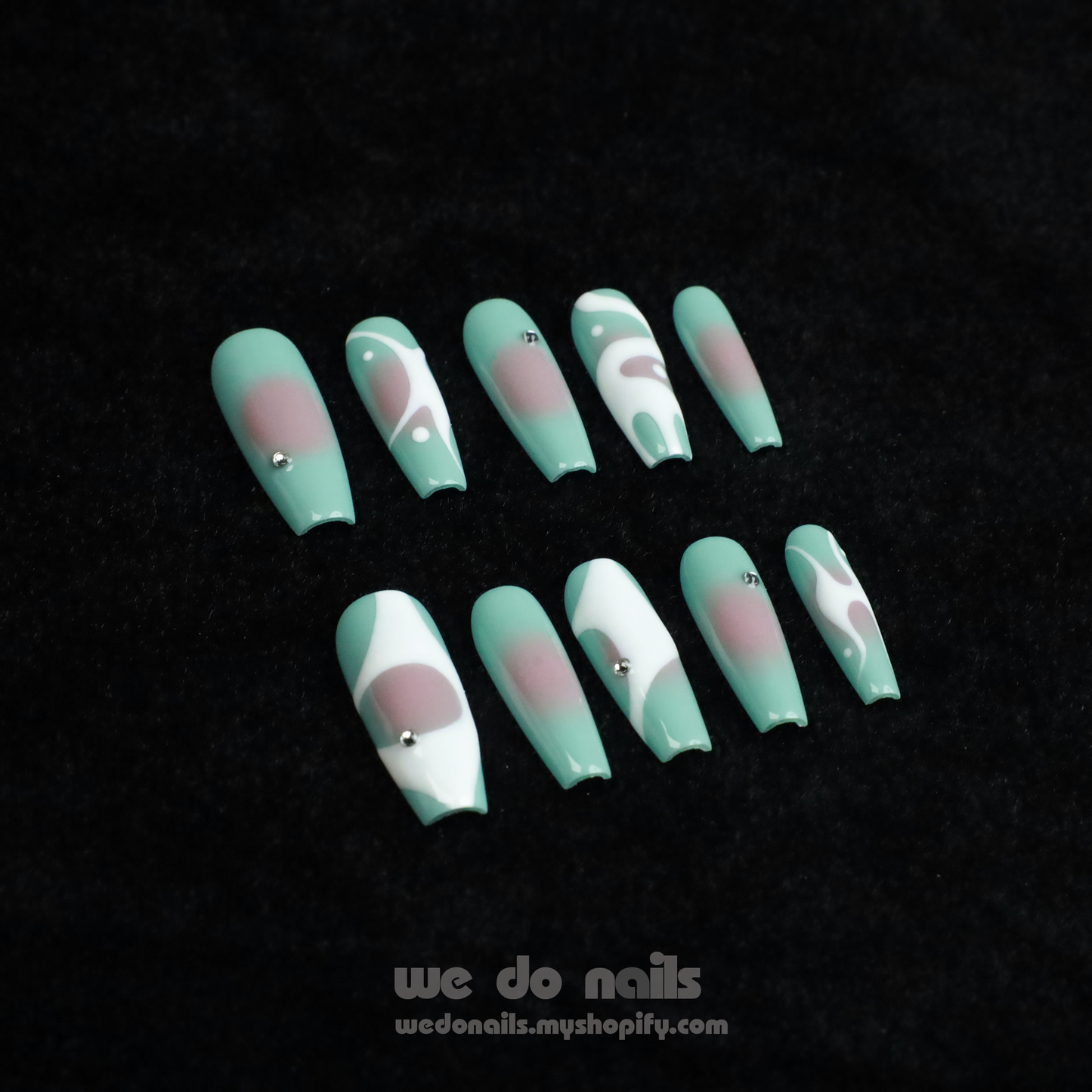 Hands with long, angular green nails pose gracefully against a black background, showcasing a modern and stylish design. The glossy finish and rings add flair, elegantly highlighting the allure of Spilled Milk Press-On Nails by We Do Nails.