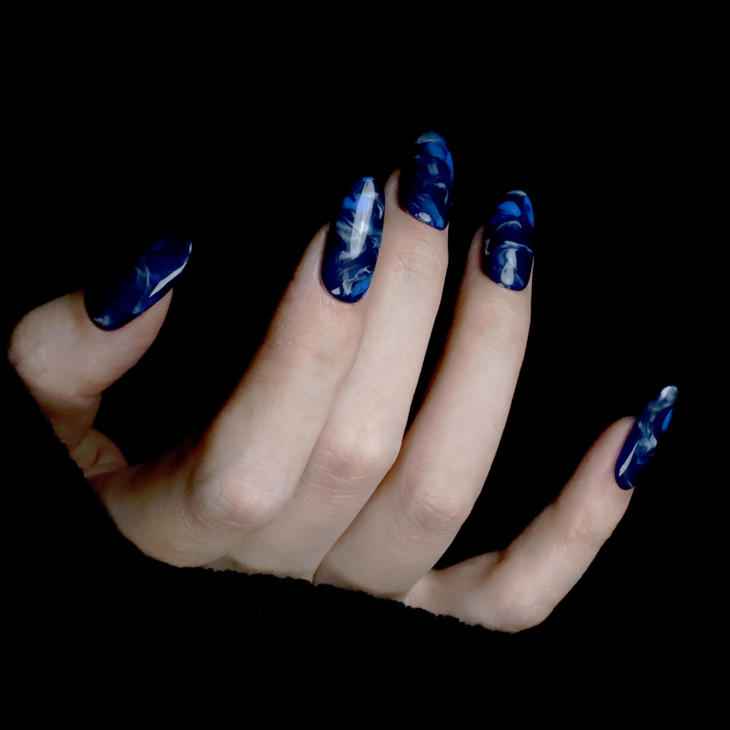 Hands display the Elegant Blue Marble Press-On Nails from We Do Nails, featuring glossy long oval shapes with a blue marble effect. A black ribbed sweater against a black background highlights these striking nails.