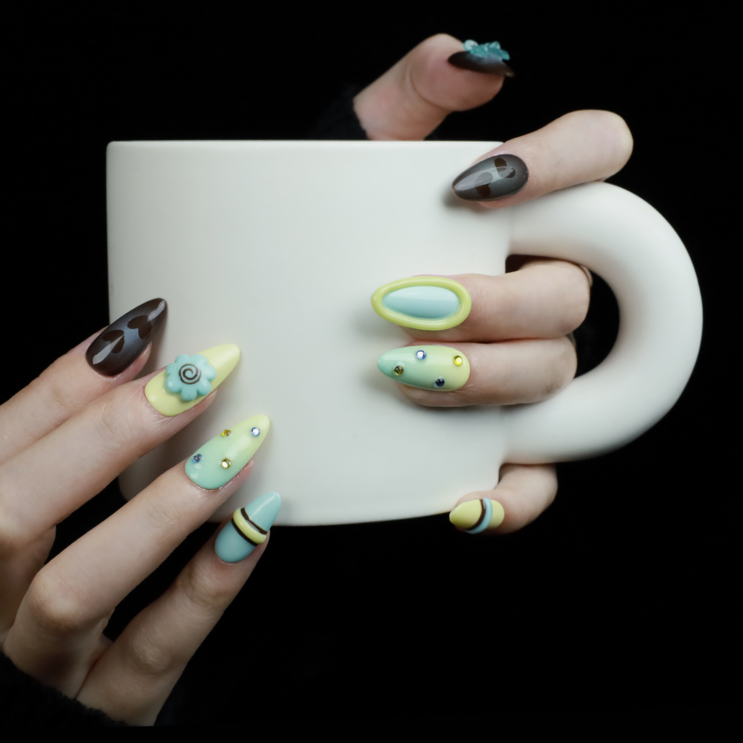A hand showcases Elegant Edge Press-On Nails by We Do Nails from the Chic Charm Collection against a black background, with designs in brown and blue gem, camo, light yellow with stripes, pale blue with rhinestones, and yellow bordered in lime green; a sleek black sleeve adorns the wrist.