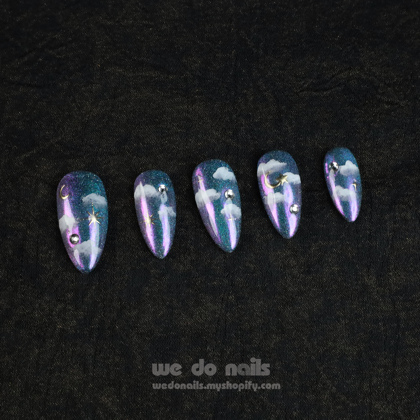 A hand showcases Starry Night Press-On Nails with long, glittery tips featuring a cloud and starry night design adorned with small gems. The person wears a black sleeve. Text below reads: We Do Nails with a website link.