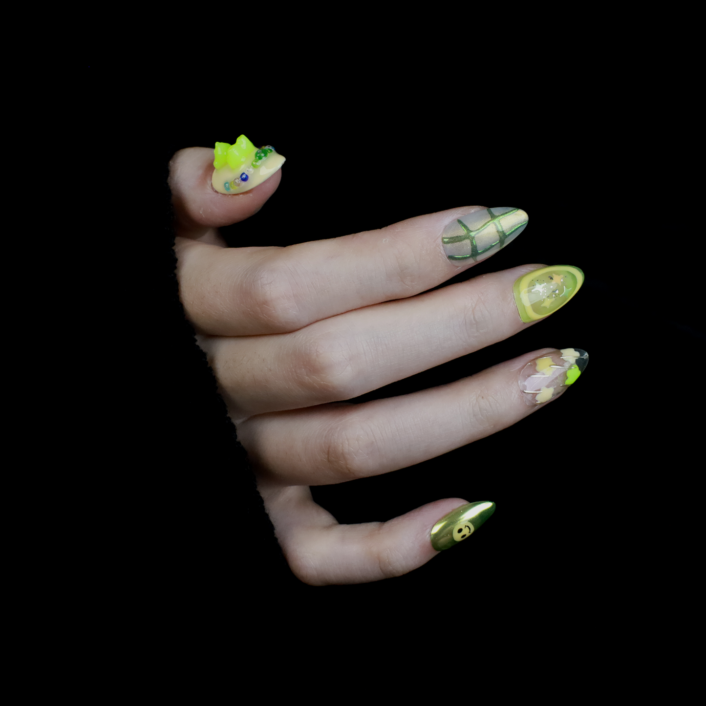 A hand holding a black mug flaunts vibrant We Do Nails Neon Slime Fantasy Press-On Nails, featuring abstract lime green, yellow, and black patterns. Spirals, dots with bead accents, and 3D smiley faces create playful art against the dark backdrop.