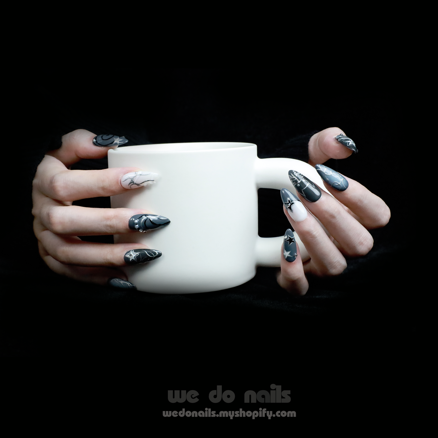 A person holds a white mug with both hands, showcasing long, pointed nails from We Do Nails Midnight Custom Press-On Nails. Star and galaxy designs exude cosmic elegance against a dark background, enhancing the vibrant celestial wonder of the nail art.