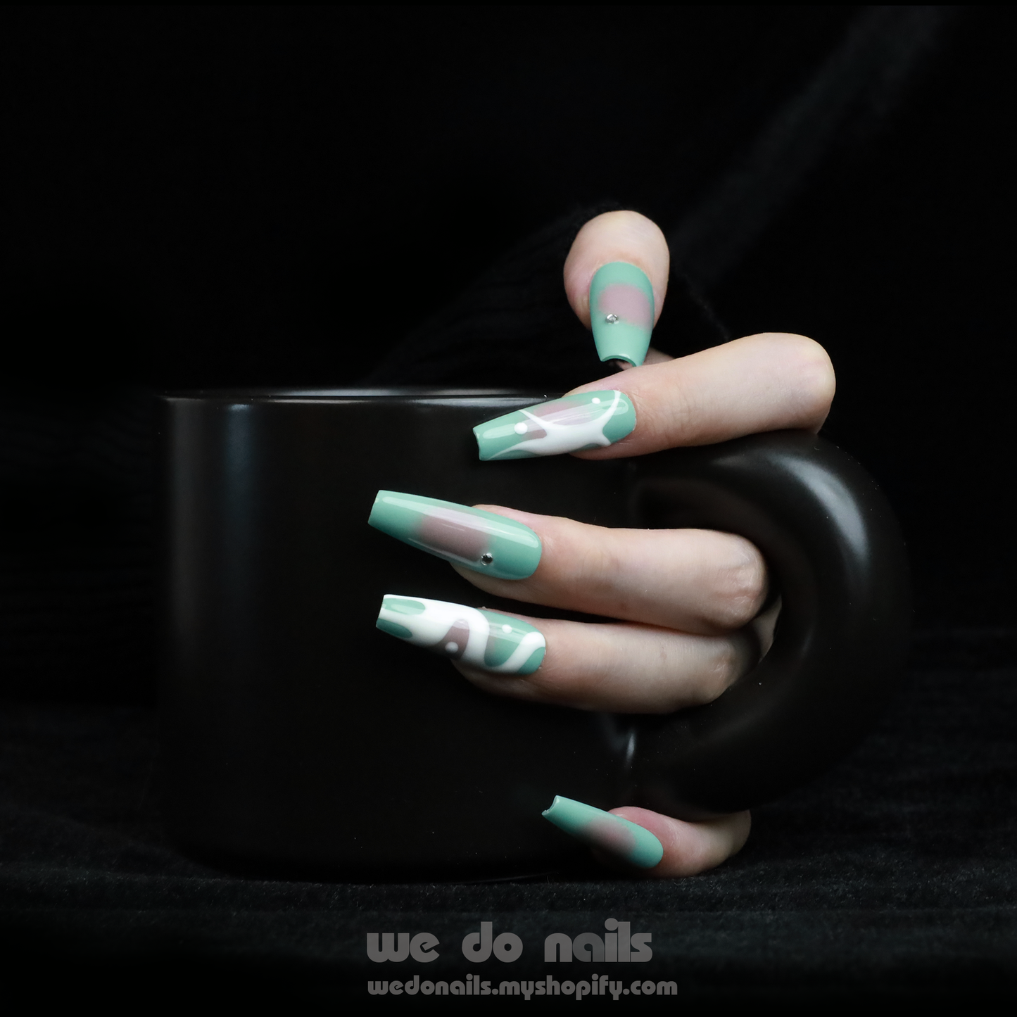 Hands with long, angular green nails pose gracefully against a black background, showcasing a modern and stylish design. The glossy finish and rings add flair, elegantly highlighting the allure of Spilled Milk Press-On Nails by We Do Nails.