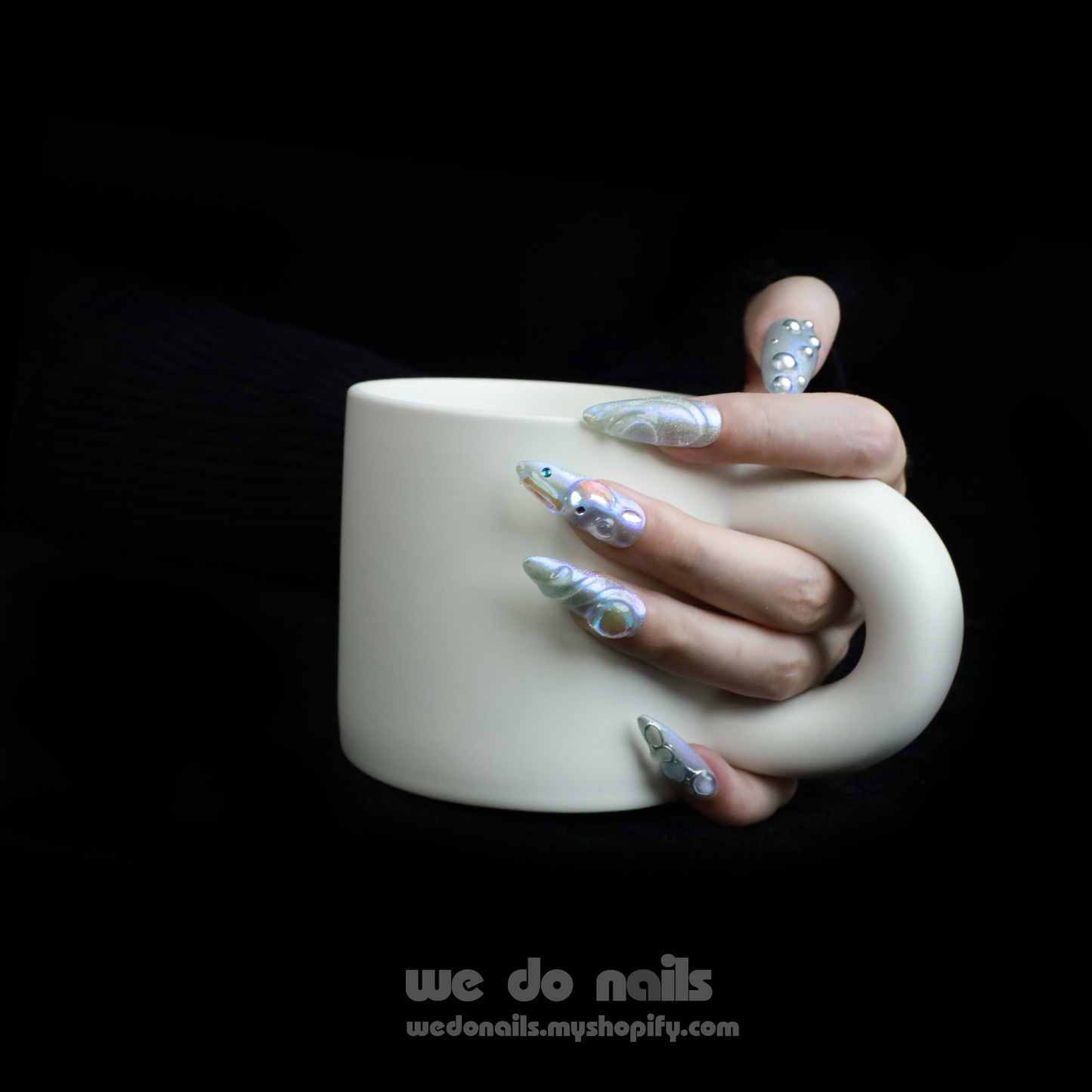 Displaying interlocked fingers adorned with We Do Nails Iridescent Bubble Press-On Nails, these long, pointed artificial nails feature a holographic silver and pearl finish, beautifully contrasted by a sleek black garment. Text reads we do nails and wedonails.myshopify.com.
