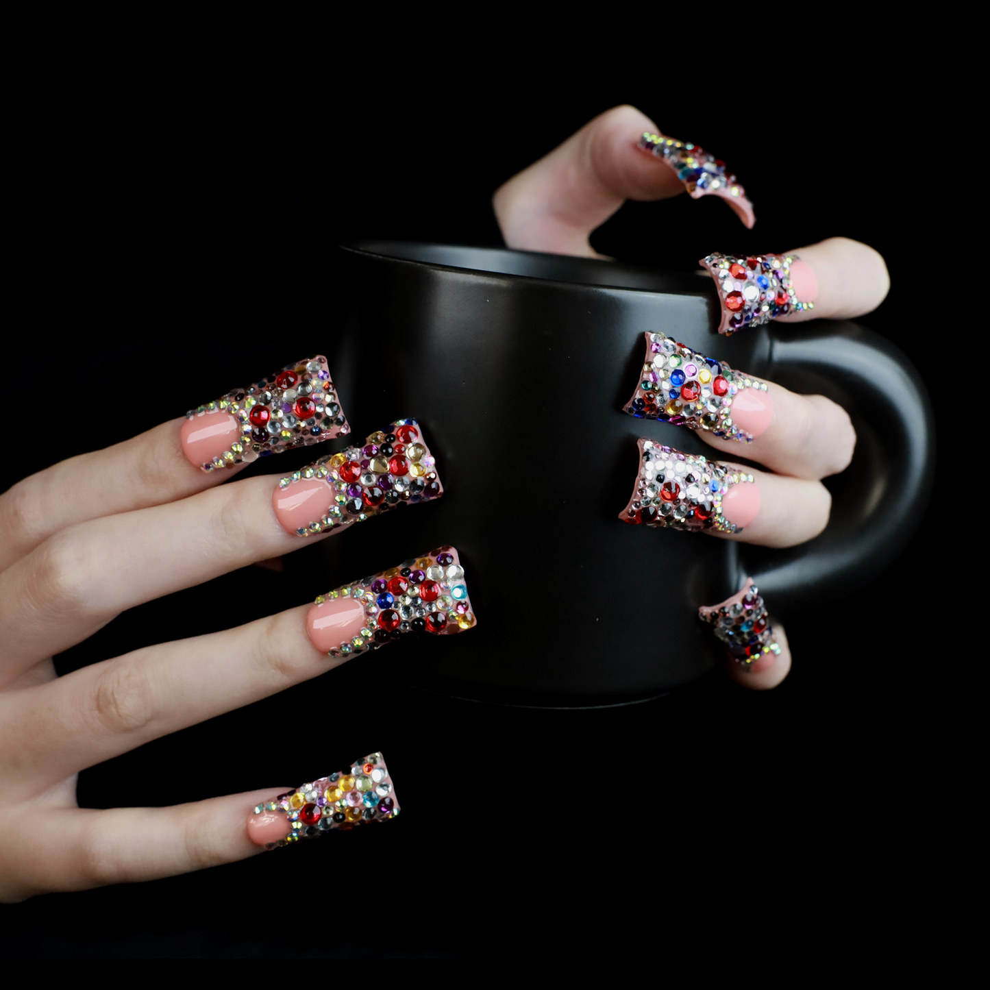 A hand flaunts Dazzling Long Duck Press-On Nails from We Do Nails, featuring a nude pink base with multicolored rhinestones against a black background. The SHIP NEXT DAY label is clearly visible in the top left corner.