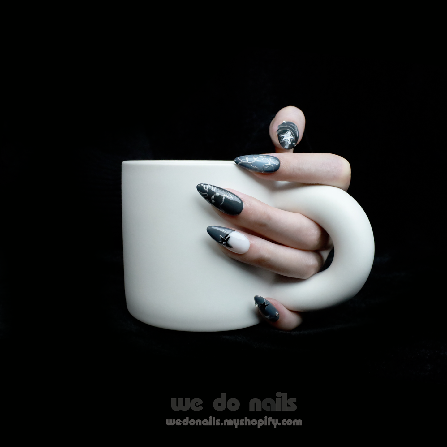 A person holds a white mug with both hands, showcasing long, pointed nails from We Do Nails Midnight Custom Press-On Nails. Star and galaxy designs exude cosmic elegance against a dark background, enhancing the vibrant celestial wonder of the nail art.