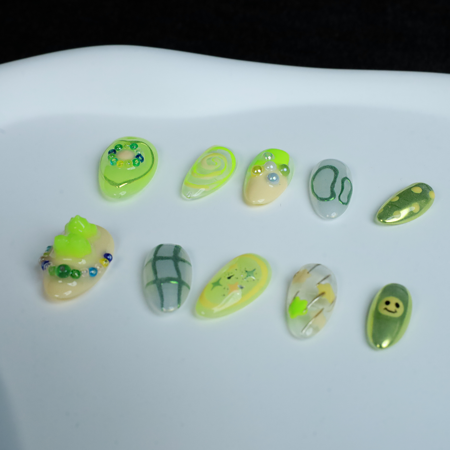 A hand holding a black mug flaunts vibrant We Do Nails Neon Slime Fantasy Press-On Nails, featuring abstract lime green, yellow, and black patterns. Spirals, dots with bead accents, and 3D smiley faces create playful art against the dark backdrop.