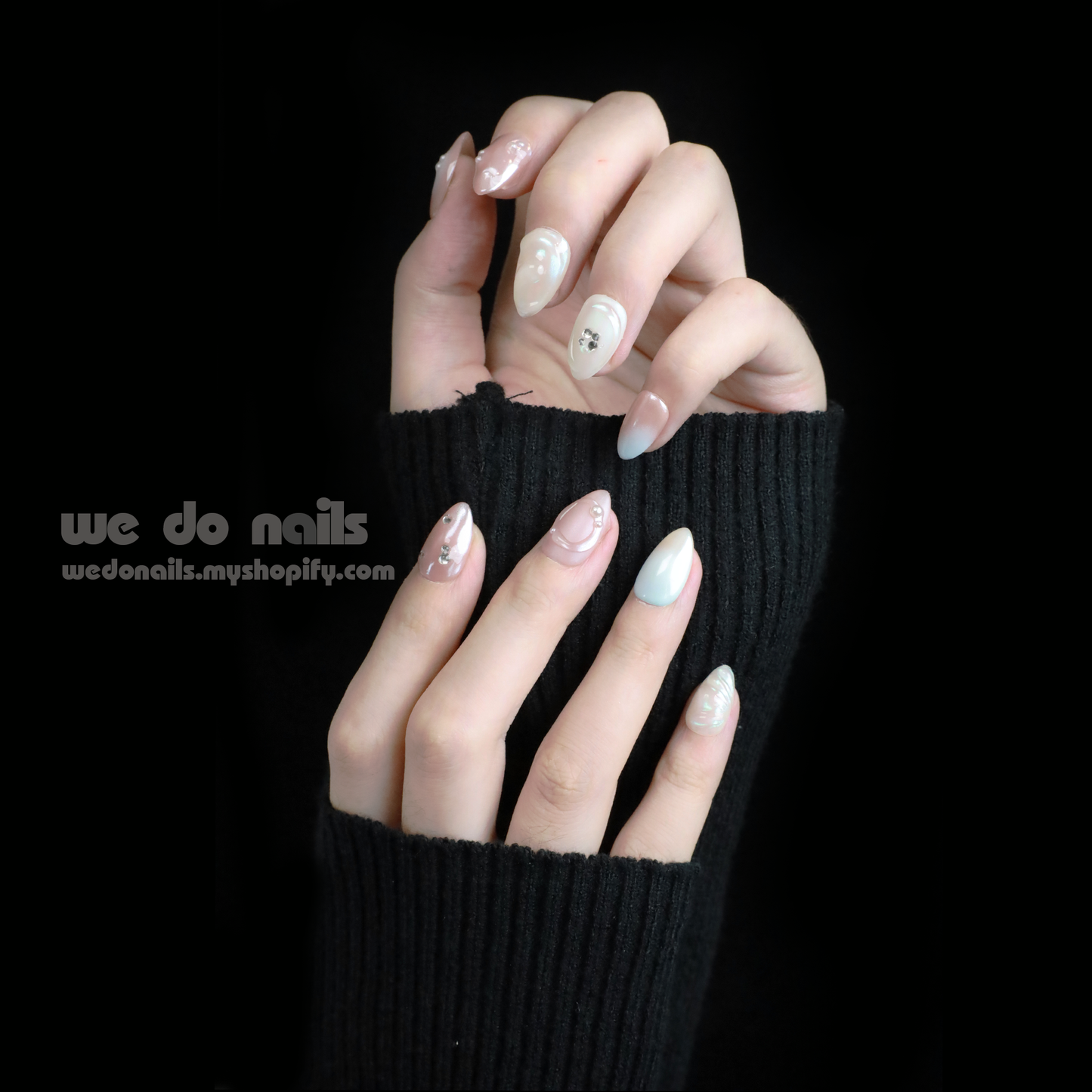 Close-up of hands with Pearl Dreams Press-On Nails by We Do Nails, showcasing manicured nails in small patterns and neutral colors. Subtle pearlescent hues add elegance. Fingers are gently intertwined, wearing a black long-sleeve sweater. We Do Nails text visible on the left.
