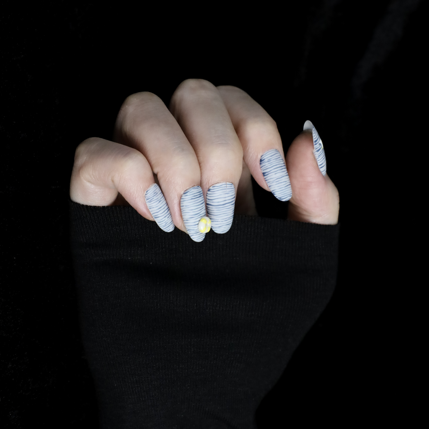 A hand with long nails showcases We Do Nails Nautical Navy Blue Stripes press-on nails, featuring a nautical blue and black wavy design. One nail is adorned with a 3D yellow flower, another with a small gem. A black sleeve adds elegance to the hand-painted look.