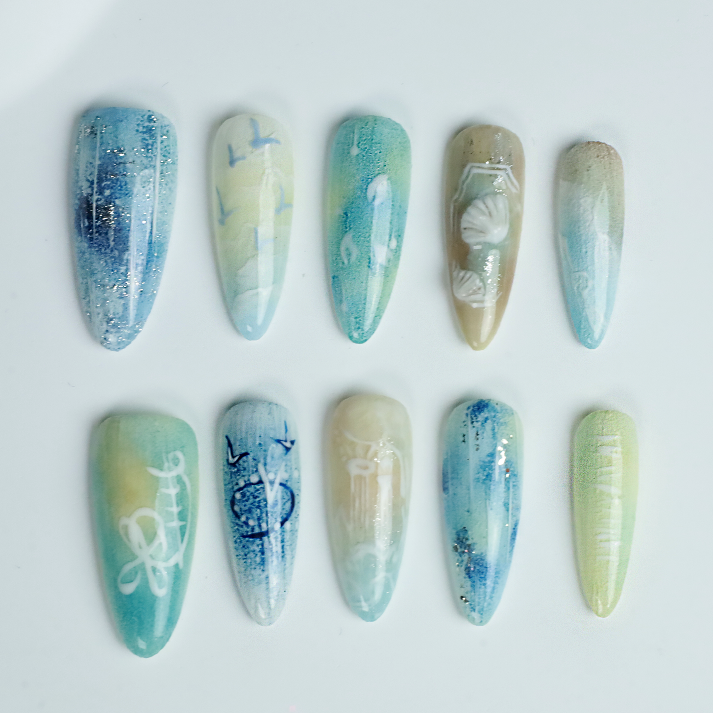 A hand flaunts the elegant Ocean-Inspired Press-On Nails by We Do Nails, featuring long, pointed shapes with ocean-themed designs. The set showcases ocean breeze colors, seashell art, and wave patterns against a dark background to highlight its intricate beauty.