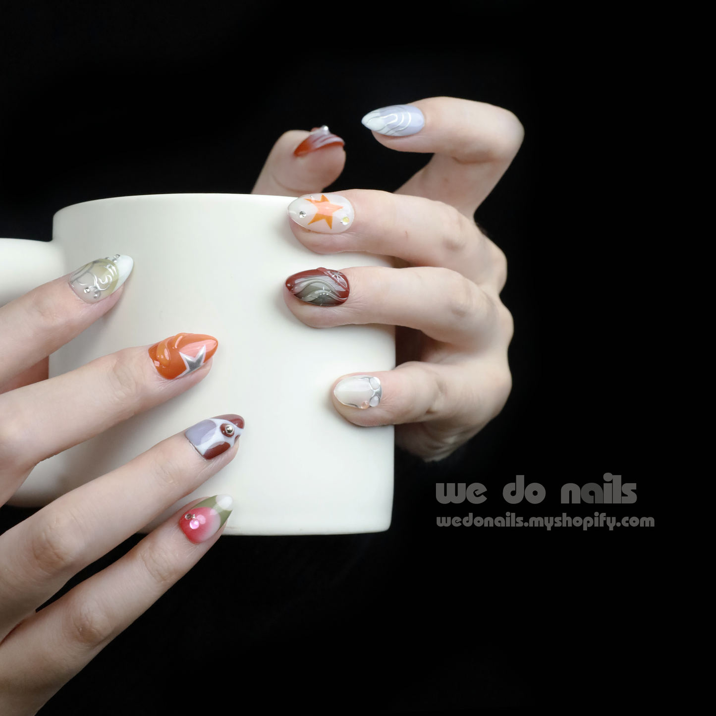 A hand displays a salon-quality manicure with Galaxy Glam Press-On Nails by We Do Nails, featuring an intricate orange and silver geometric design. The cosmic vibe shines against the dark backdrop, while SHIP NEXT DAY stands out in a pink rectangle in the upper left.