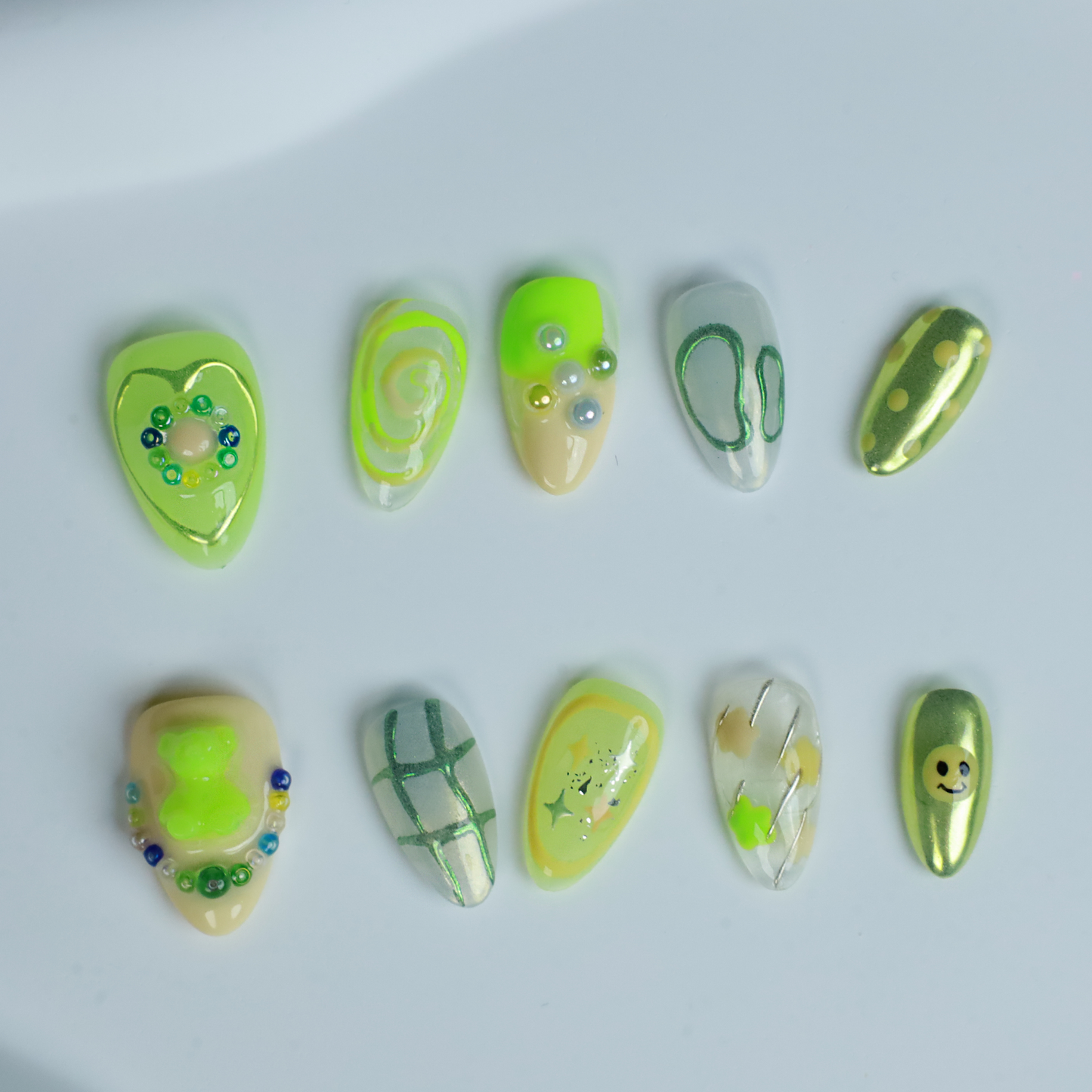 A hand holding a black mug flaunts vibrant We Do Nails Neon Slime Fantasy Press-On Nails, featuring abstract lime green, yellow, and black patterns. Spirals, dots with bead accents, and 3D smiley faces create playful art against the dark backdrop.