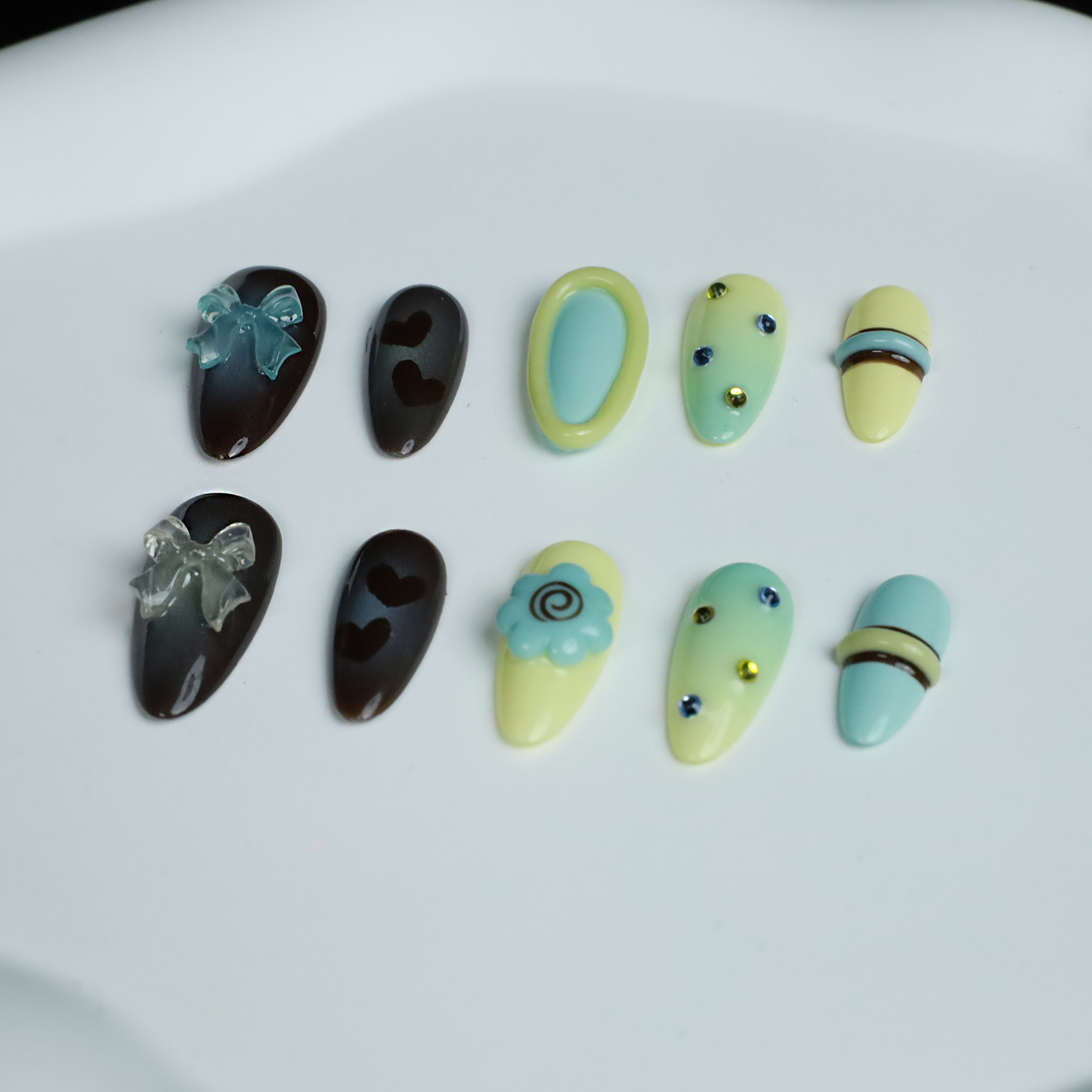 A hand showcases Elegant Edge Press-On Nails by We Do Nails from the Chic Charm Collection against a black background, with designs in brown and blue gem, camo, light yellow with stripes, pale blue with rhinestones, and yellow bordered in lime green; a sleek black sleeve adorns the wrist.