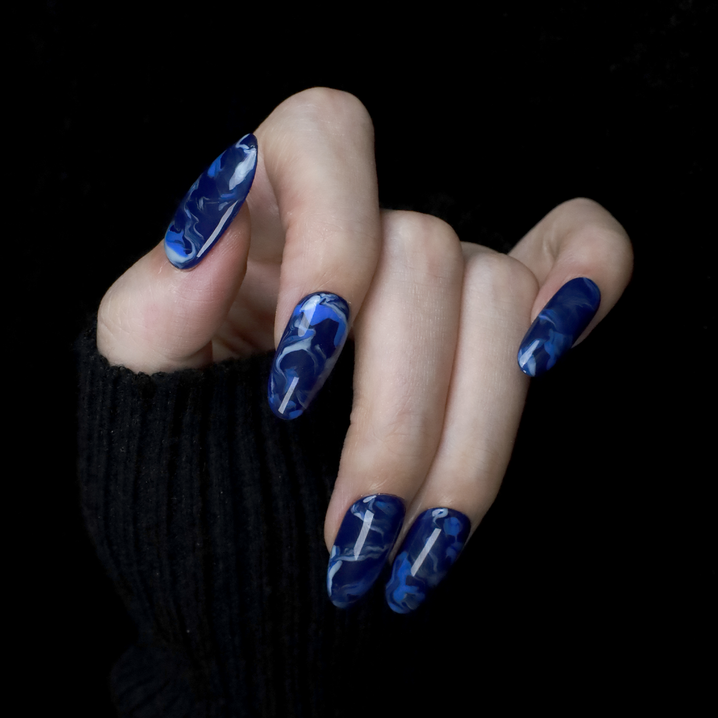 Hands display the Elegant Blue Marble Press-On Nails from We Do Nails, featuring glossy long oval shapes with a blue marble effect. A black ribbed sweater against a black background highlights these striking nails.