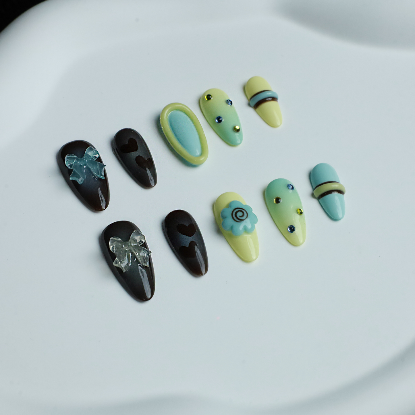 A hand showcases Elegant Edge Press-On Nails by We Do Nails from the Chic Charm Collection against a black background, with designs in brown and blue gem, camo, light yellow with stripes, pale blue with rhinestones, and yellow bordered in lime green; a sleek black sleeve adorns the wrist.