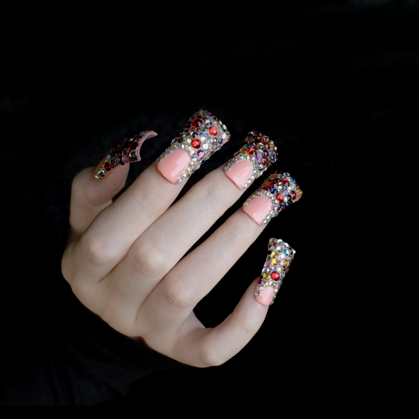 A hand flaunts Dazzling Long Duck Press-On Nails from We Do Nails, featuring a nude pink base with multicolored rhinestones against a black background. The SHIP NEXT DAY label is clearly visible in the top left corner.