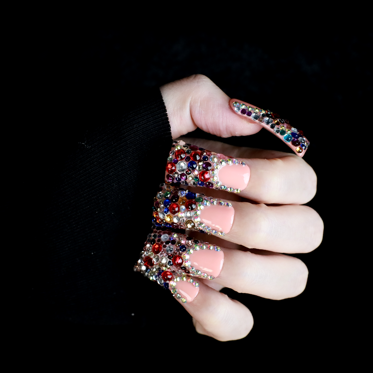 A hand flaunts Dazzling Long Duck Press-On Nails from We Do Nails, featuring a nude pink base with multicolored rhinestones against a black background. The SHIP NEXT DAY label is clearly visible in the top left corner.