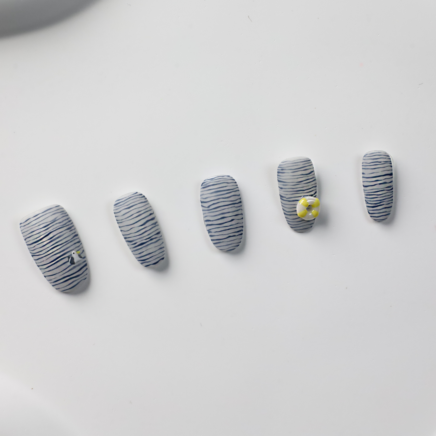A hand with long nails showcases We Do Nails Nautical Navy Blue Stripes press-on nails, featuring a nautical blue and black wavy design. One nail is adorned with a 3D yellow flower, another with a small gem. A black sleeve adds elegance to the hand-painted look.