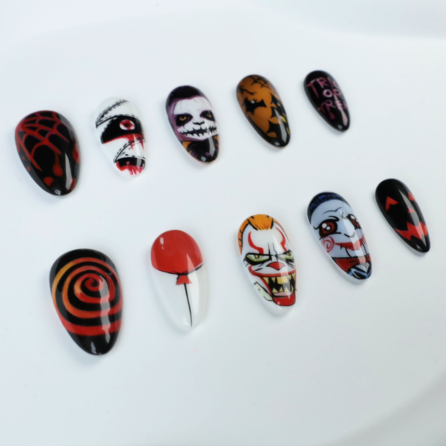 A close-up of two hands shows off the eerie Spooky Nightmares Halloween Press-On Nails by We Do Nails, featuring hand-painted designs like clowns, a pumpkin, and abstract art on a black background while holding a large white mug.