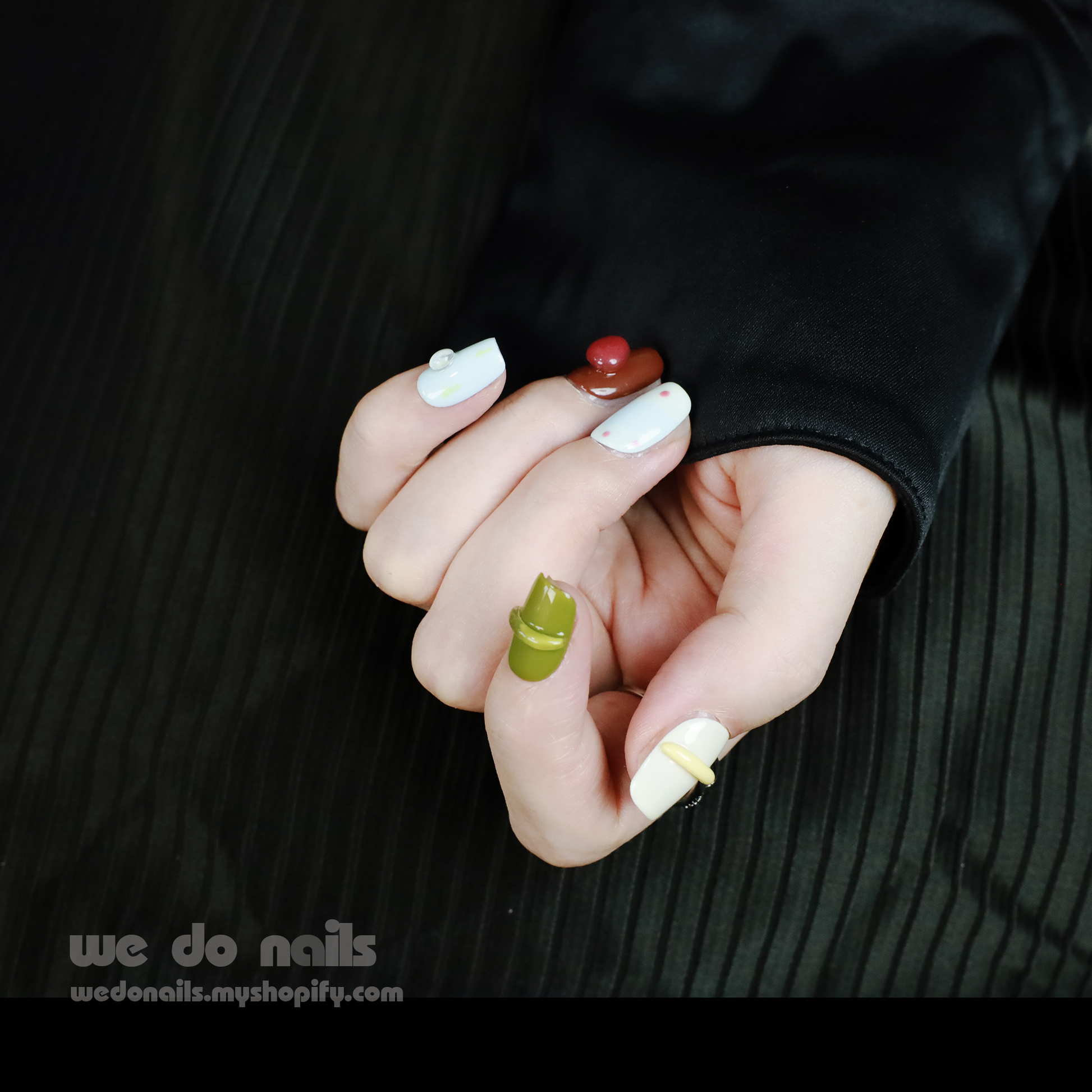 Close-up of colorful hands against a dark background, featuring We Do Nails Cute Abstract Art Press-On Nails with vibrant patterns like stripes and polka dots. Some nails have 3D finishes and embellishments, complemented by several rings on the persons fingers.
