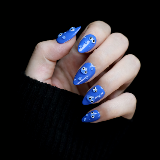 A hand with We Do Nails Cute Blue Monster Press-On Nails set features long, almond-shaped, bright blue nails adorned with playful cartoon faces and 3D googly eyes. The hand in a black sweater is striking against the dark background.