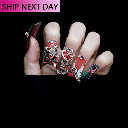A hand displays Glam Jungle Luxe 3D Long Duck Press-On Nails by We Do Nails, featuring ornate black and red designs with rhinestone gemstones and skulls. The words SHIP NEXT DAY appear in the top-left corner, highlighting their gothic style against a dark background.