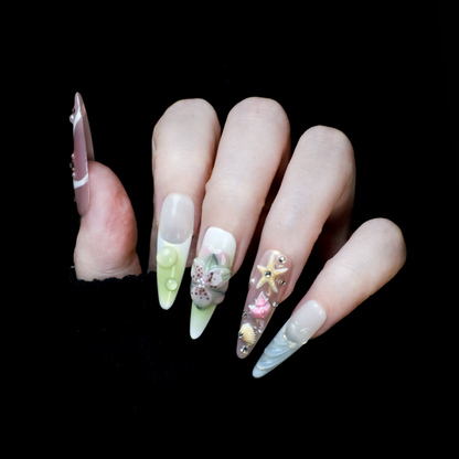 A hand flaunts We Do Nails Floral Blush Pearl Press-On Nails: green with pearls, pink with a butterfly, clear with gold accents, blue with a wave pattern, and a pink-white thumb—all set against a black background for an enchanting look.