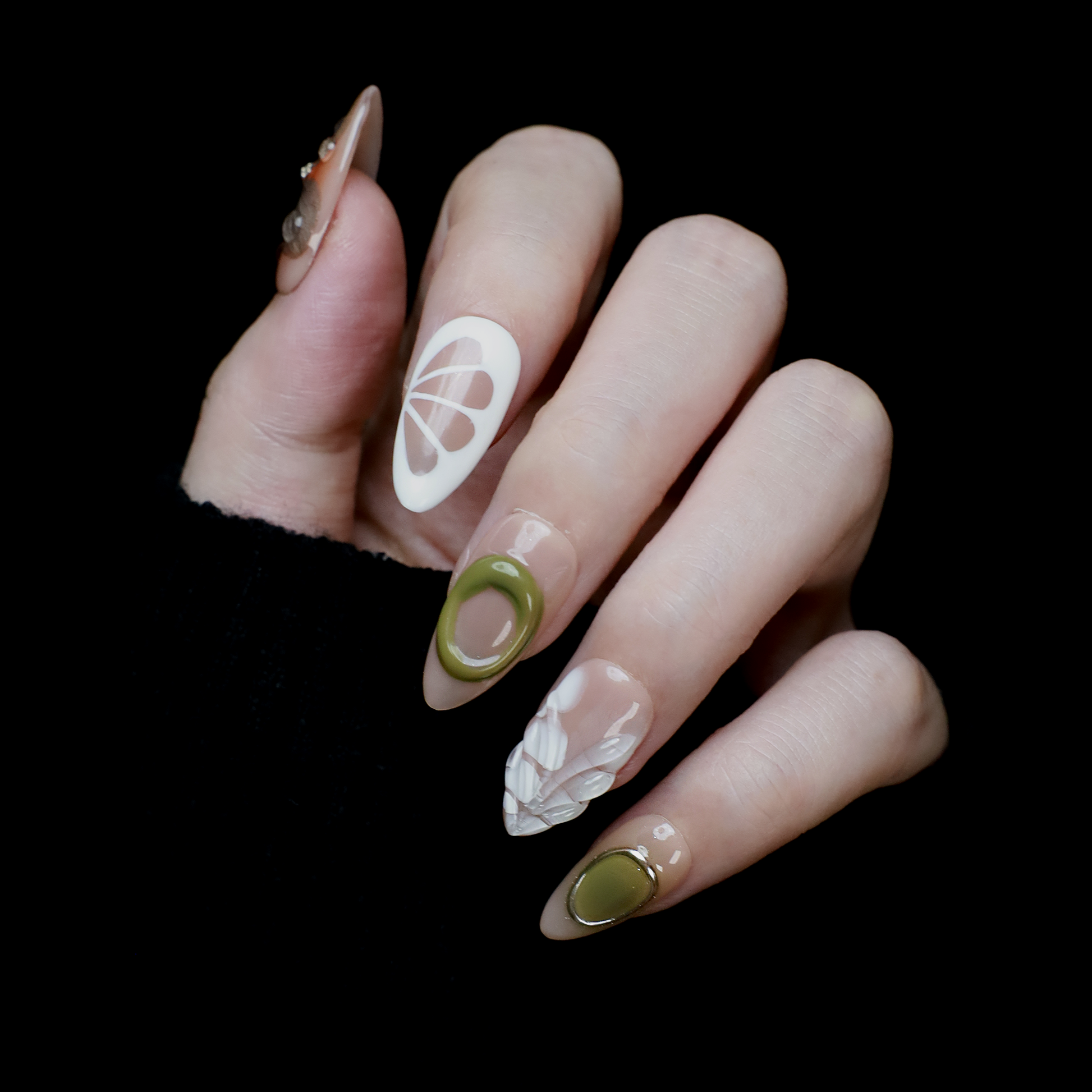 A hand in a black sleeve displays the Leaf and Organic design Press-On Nails by We Do Nails, featuring long acrylic nails with white abstract patterns, green accents, and clear 3D embellishments that create a nature-inspired look against the dark background.