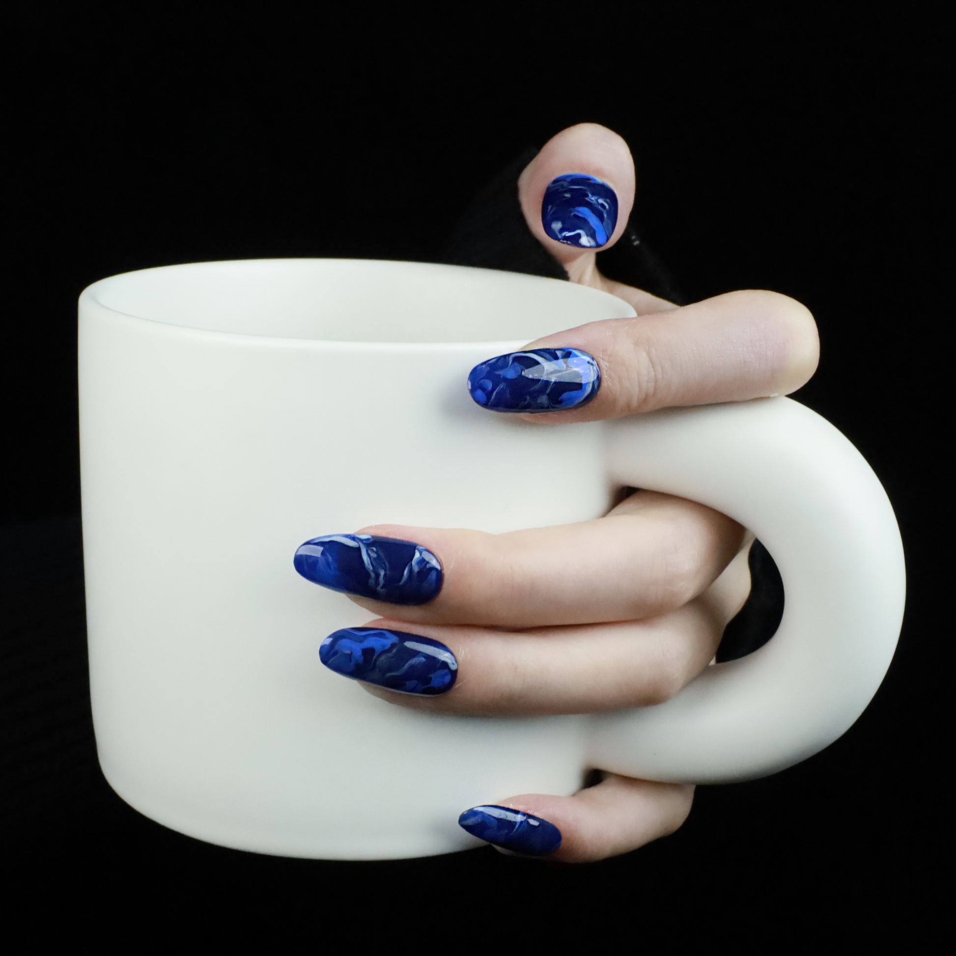 Hands display the Elegant Blue Marble Press-On Nails from We Do Nails, featuring glossy long oval shapes with a blue marble effect. A black ribbed sweater against a black background highlights these striking nails.