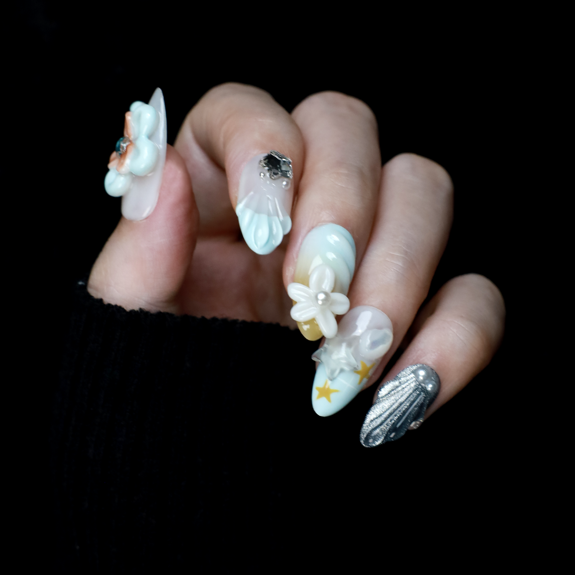 A hand with long, intricately decorated nails exhibits ethereal designs of blue and white swirls, 3D floral accents, pearls, and a metallic silver textured nail against a black background. This look perfectly captures the elegance of We Do Nails Celestial Blossom Press On Nails.