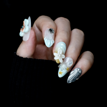 A hand with long, intricately decorated nails exhibits ethereal designs of blue and white swirls, 3D floral accents, pearls, and a metallic silver textured nail against a black background. This look perfectly captures the elegance of We Do Nails Celestial Blossom Press On Nails.