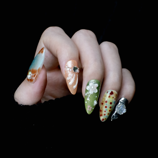 A hand showcases the Fantasy Garden Press-On Nails by We Do Nails against a black background. The nails feature designs of a koi fish with a pearl, peach with rhinestones, 3D green flowers, green with red floral dots, and metallic silver with a crystal accent.