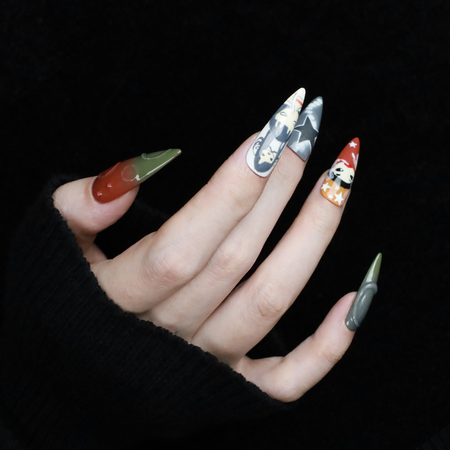 A hand with long, pointed Japanese-Inspired Artistic Press-On Nails by We Do Nails showcases camo patterns in green, red, and gray. Combining kawaii aesthetic and artistic motifs that suggest anime nail art, the hand is partially covered by a black sleeve.