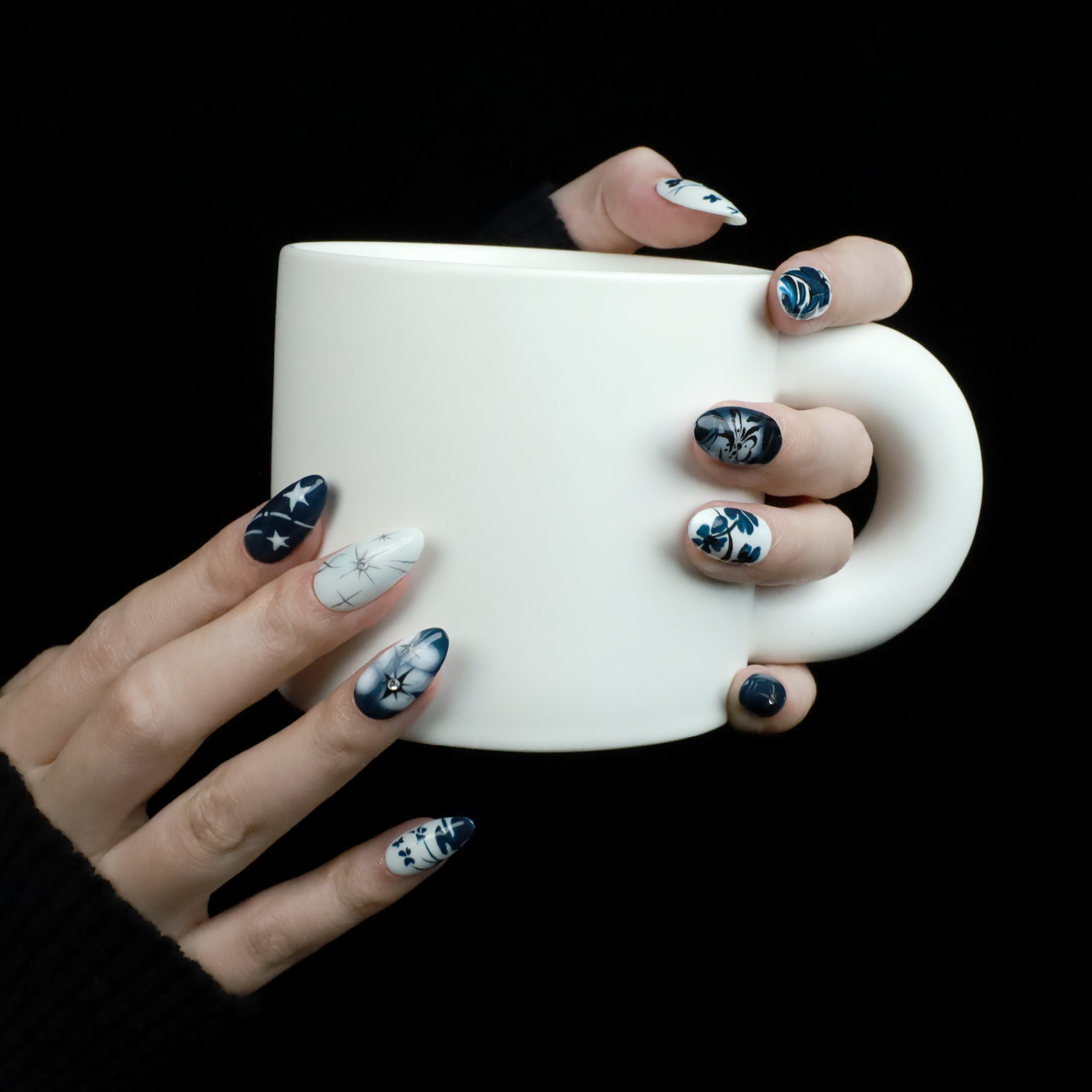 Hands adorned with We Do Nails Blue & White Porcelain Inspired Nails, showcasing long, almond-shaped designs with intricate traditional floral and abstract patterns. The nails stand out against black sleeves and a black background, reflecting the elegance of Chinese porcelain.