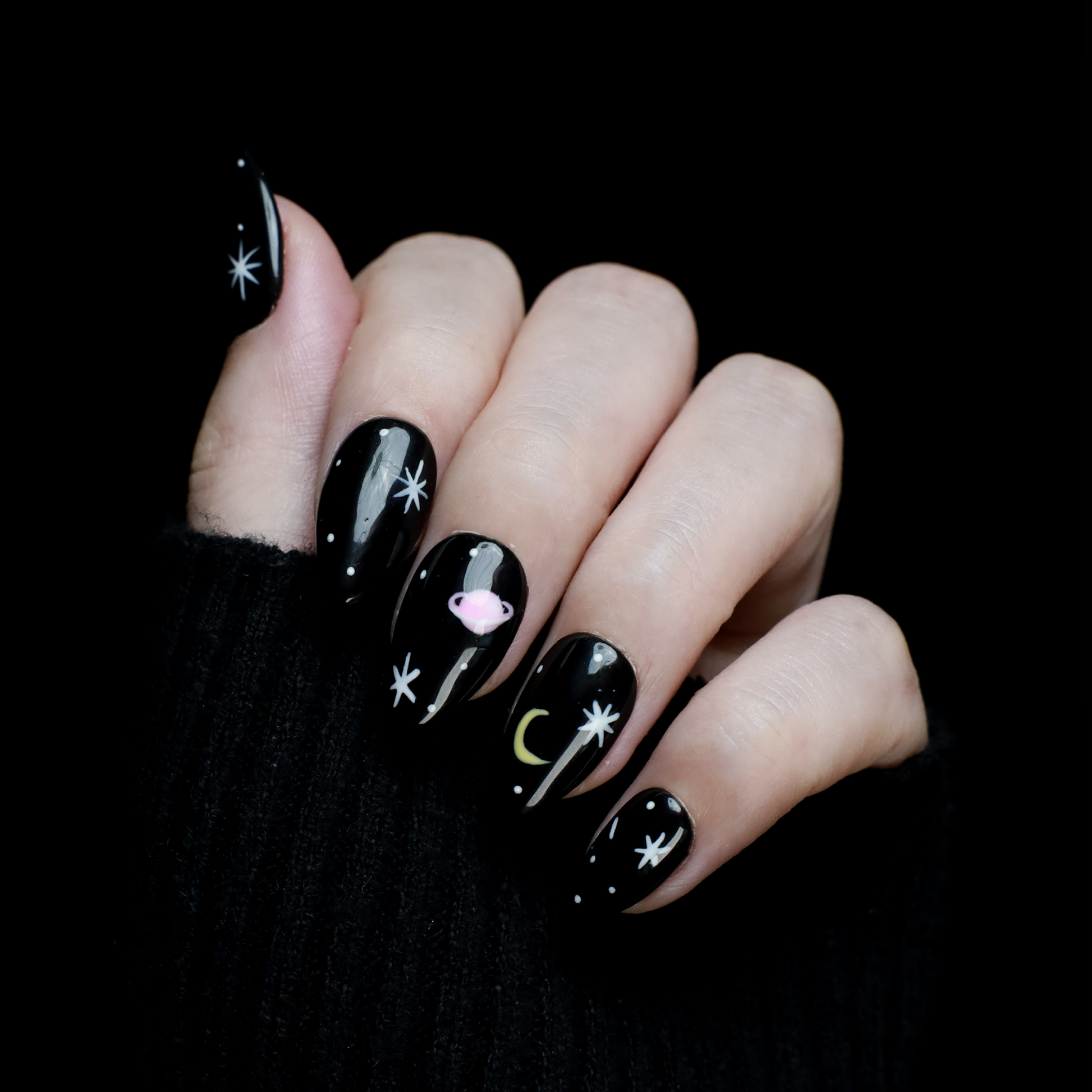 Hands decorated with Galactic Night Sky Press On Nails by We Do Nails, featuring stars, moons, and planets, hold a plain white mug against a dark backdrop.