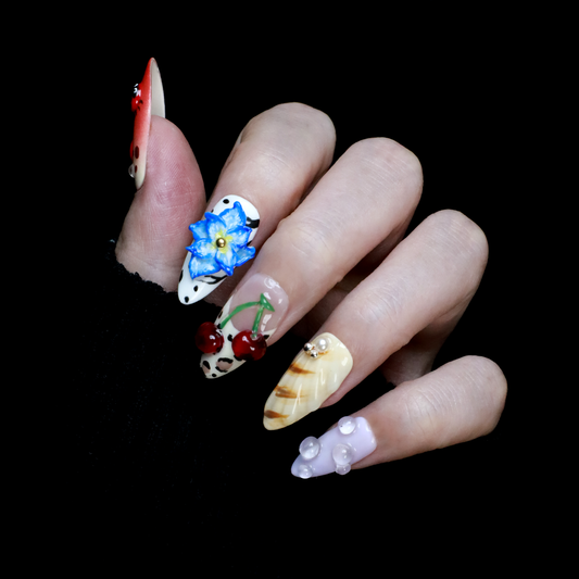 The Garden Blooms Press-On Nails by We Do Nails feature a whimsical garden theme with long, decorative nails, including a blue flower, cherries, red and white patterns, pearls, and stripes. Set against a black background, the hand is partially covered by a black sleeve.