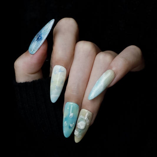 A hand flaunts the elegant Ocean-Inspired Press-On Nails by We Do Nails, featuring long, pointed shapes with ocean-themed designs. The set showcases ocean breeze colors, seashell art, and wave patterns against a dark background to highlight its intricate beauty.