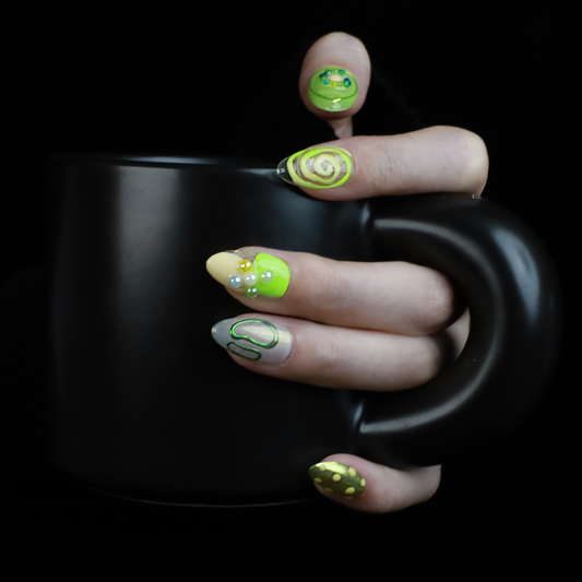 A hand holding a black mug flaunts vibrant We Do Nails Neon Slime Fantasy Press-On Nails, featuring abstract lime green, yellow, and black patterns. Spirals, dots with bead accents, and 3D smiley faces create playful art against the dark backdrop.