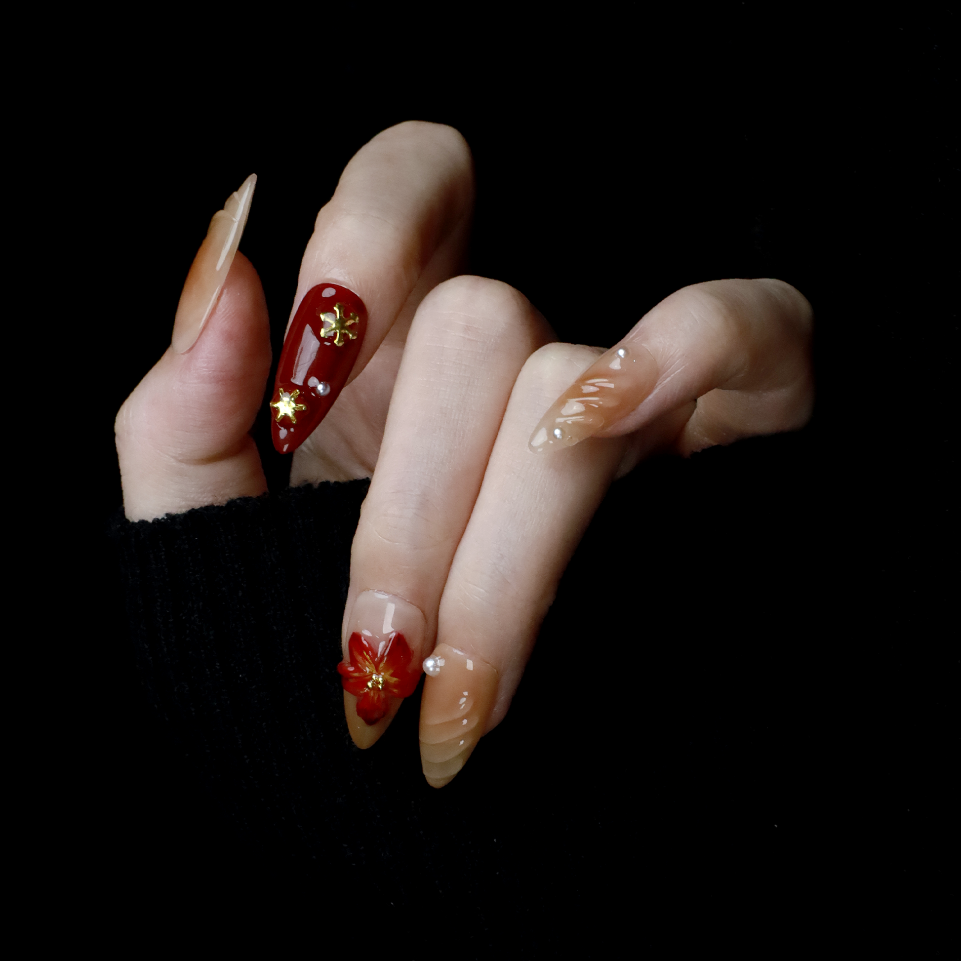 A pair of hands adorned with Elegant Floral Red and Beige Press-On Nails by We Do Nails, featuring intricate red flowers and pearl-gold accents, holds a large black mug. The elongated, almond-shaped nails boast a glossy finish with 3D floral designs against the black background.